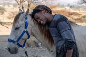 Tajebu, wearing a dark and light blue sweater, hugs the side of Bora, a white horse in blue harnessing.
