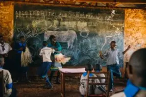 A child stands at the front of a Donkey Care Club class, while others draw donkeys on the chalk board.