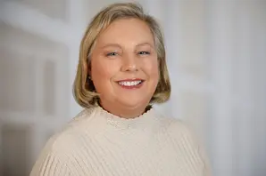 Lin McGarry, smiles while standing against a white background and wearing a white turtle neck jumper.