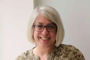 Nicola Chevis stands in front of a white background while smiling and wearing a green and white blouse.