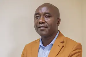 Raphael Kinoti wearing a yellow jacket and blue shirt, stands with his arms folded and smiling against a beige background