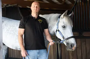 Sam Dracott stands, wearing a black t-shirt and blue jeans, in front of a white horse.