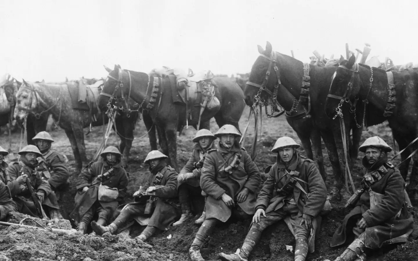 A row of soldiers sit in front of their horses in the trenches during World War One, in a black and white photo.