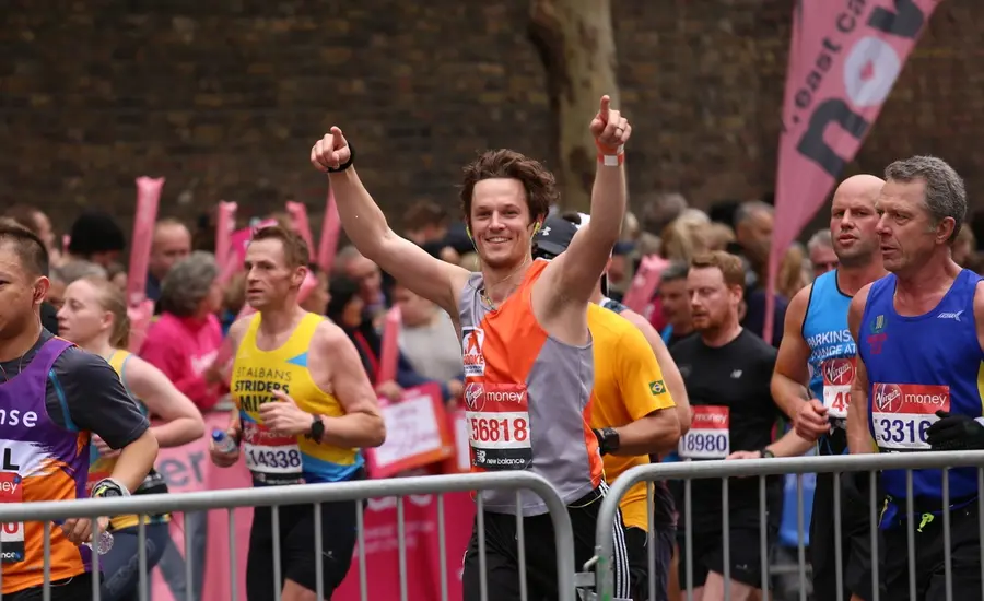 A person running in a marathon, wearing a grey and orange vest and black shorts and waving their arms in the air