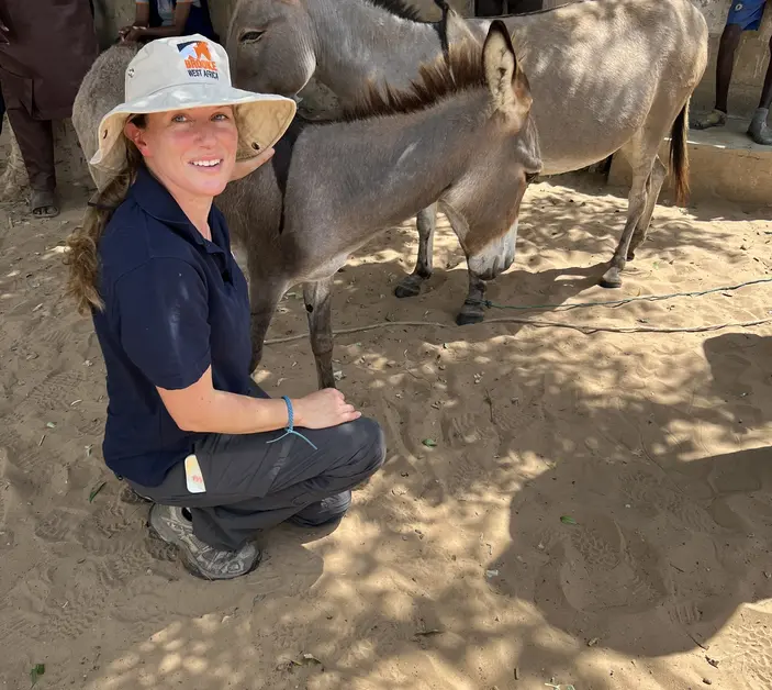 Dr Laura Skippen, wearing a navy blue t-shirt and Brooke sun hat, crouches down and makes friends with a donkey foal