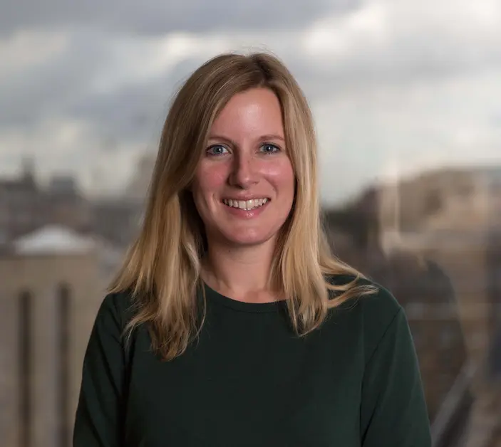 Brooke's Dr Laura Skippen stands in front of a city skyline wearing a charcoal jumper