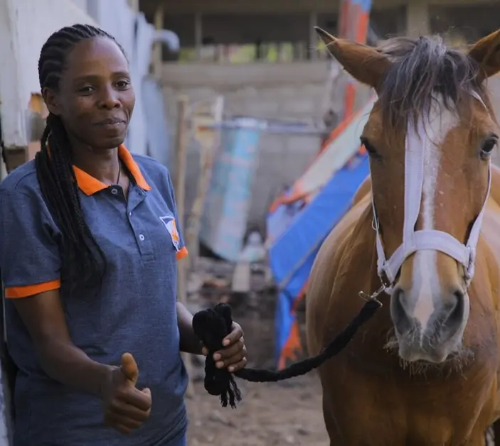 A woman in a blue and orange Brooke t-shirt stands next to a brown horse.