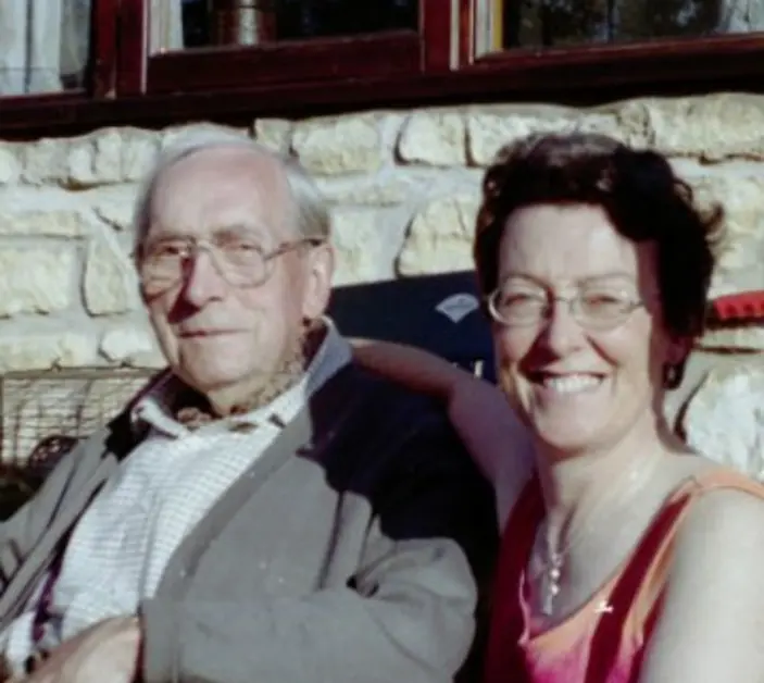 Diana, wearing a pink blouse, sits with her dad, dressed in a white shirt and grey cardigan, outside a house.