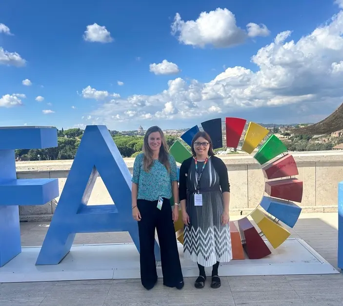 Laura and Anna stand together in front of 'FAO' in large letters against a backdrop of blue skies and countryside in Rome, Italy.