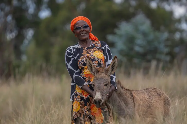 Beth stands in a field, wearing black and orange, with a donkey.